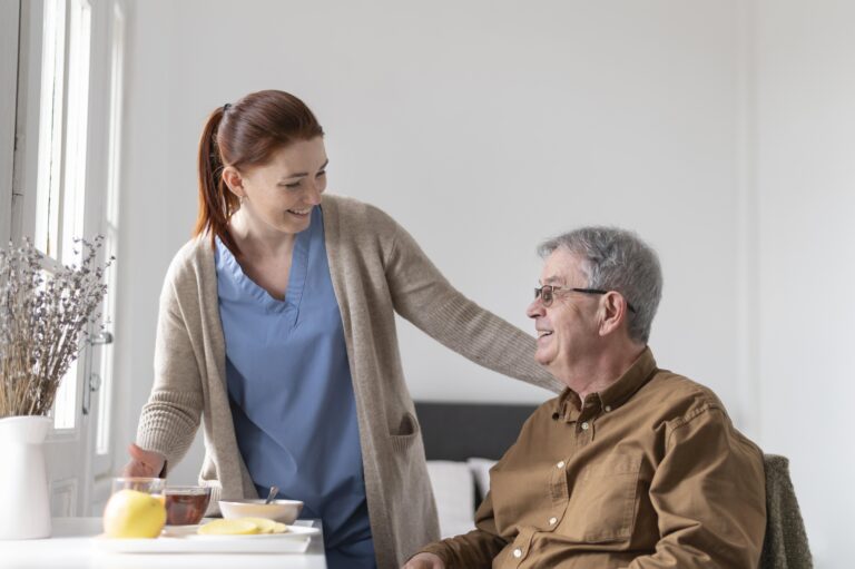 Cuidadora sorridente em jaleco azul interage com um homem idoso sentado, em um ambiente acolhedor e bem iluminado, com uma mesa ao lado contendo frutas e chá.